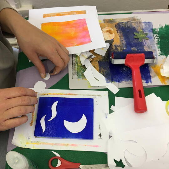 Hands placing paper shapes on Gel Printing Plate, blue inked brayer on table