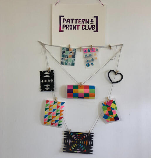 White wall with string holding tissue paper stained glass and snowflake cut designs attached with pegs. Pattern and Print club logo at top