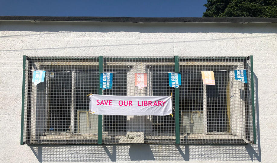 Bright neon pink text on white fabric saying 'save our library' hung on window covers
